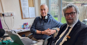 Two men in the recording studio. The mayor is on the right and the Recordist is on the left with headphones around his neck.